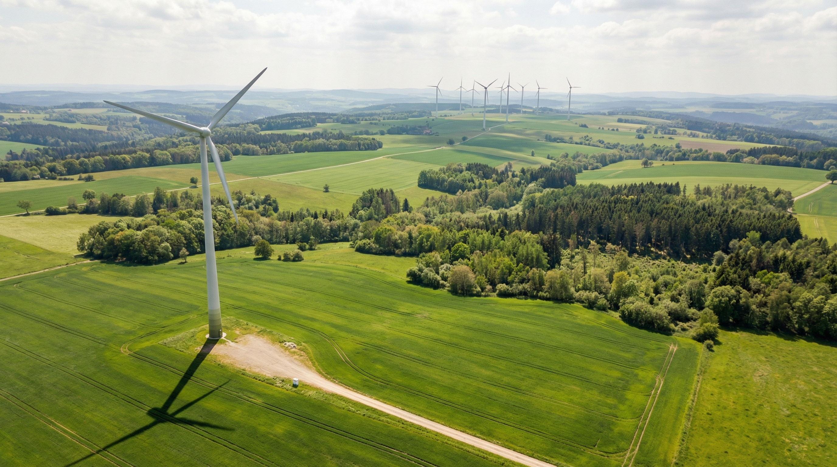 Luftaufnahme einer grünen Landschaft mit Feldern und Waldflächen. Im Vordergrund steht ein einzelnes Windrad, weitere Windräder sind in der Ferne zu sehen. Der Himmel ist leicht bewölkt und sonnig.