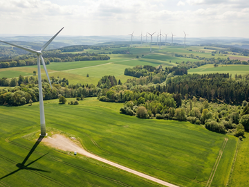 Luftaufnahme einer grünen Landschaft mit Windrädern Luftaufnahme einer grünen Landschaft mit Feldern und Waldflächen. Im Vordergrund steht ein einzelnes Windrad, weitere Windräder sind in der Ferne zu sehen. Der Himmel ist leicht bewölkt und sonnig.