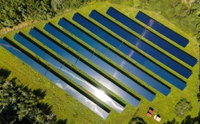 Solarpark Schkopau von oben Das Bild zeigt eine Luftaufnahme eines Solarparks mit mehreren Reihen von Solarmodulen, die auf einer grasbewachsenen Fläche installiert sind. Die Solarpaneele sind in parallelen Linien angeordnet. Auf einem der Module spiegelt sich das Sonnenlicht.