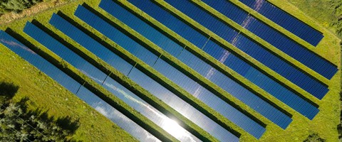 Solarpark Schkopau von oben Das Bild zeigt eine Luftaufnahme eines Solarparks mit mehreren Reihen von Solarmodulen, die auf einer grasbewachsenen Fläche installiert sind. Die Solarpaneele sind in parallelen Linien angeordnet. Auf einem der Module spiegelt sich das Sonnenlicht.