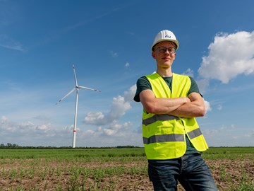 Mitarbeiter vor Feld mit Windrad Eine Person steht mit verschränkten Armen auf einem offenen Feld. Sie trägt einen weißen Schutzhelm und eine gelbe reflektierende Sicherheitsweste. Im Hintergrund befindet sich eine große Windkraftanlage mit drei Rotorblättern.