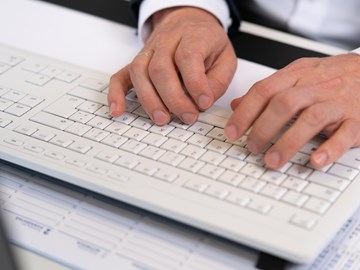 Haende auf Tastatur Das Bild zeigt eine Nahaufnahme der Hände einer Person, die auf einer weißen Tastatur tippt. Die Person trägt ein dunkles Sakko und ein weißes Hemd.