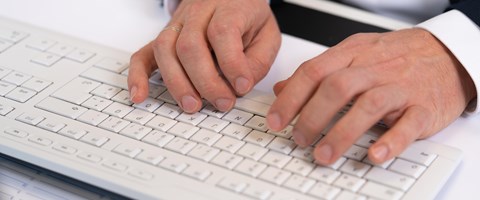 Haende auf Tastatur Das Bild zeigt eine Nahaufnahme der Hände einer Person, die auf einer weißen Tastatur tippt. Die Person trägt ein dunkles Sakko und ein weißes Hemd.