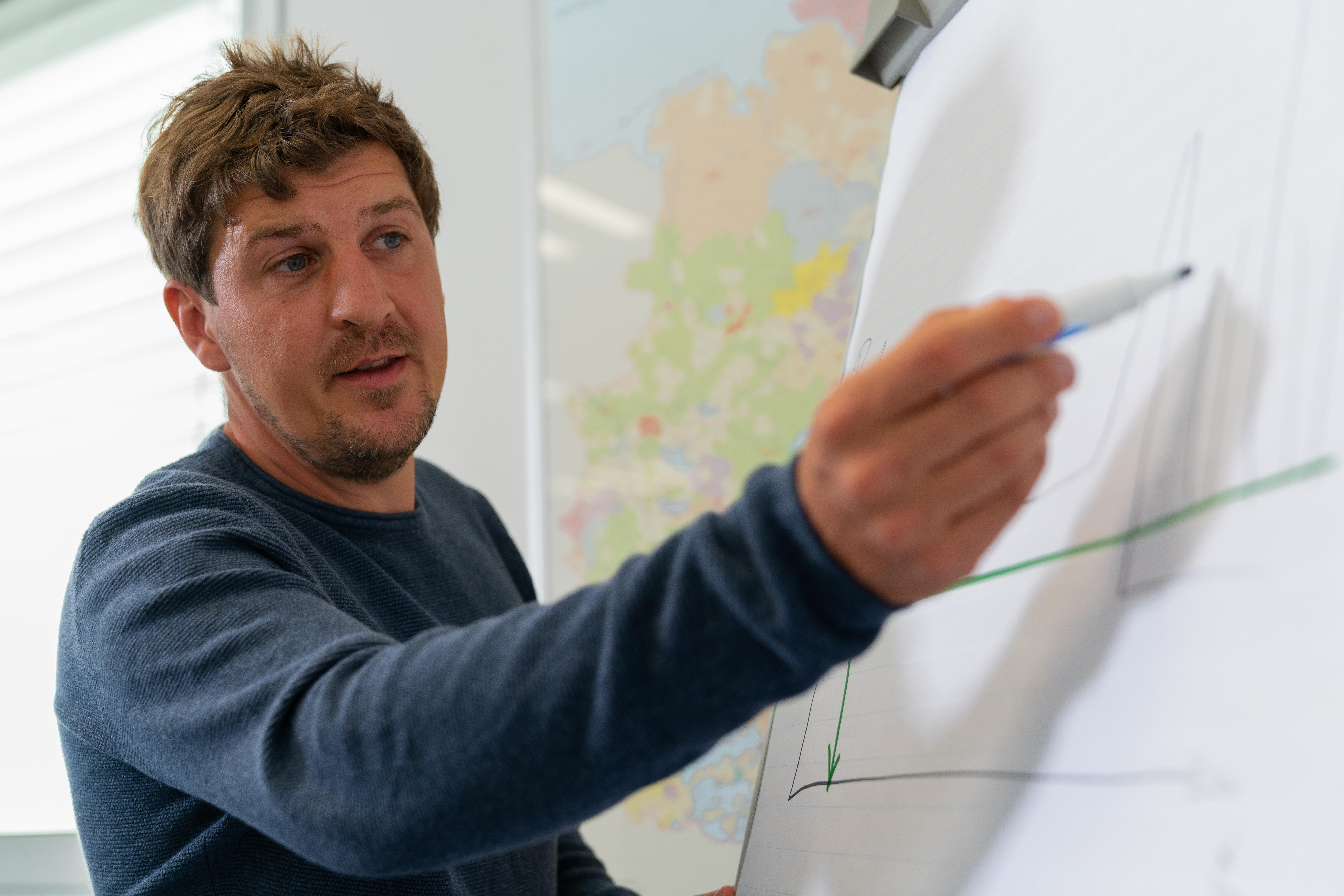 Eine Person zeigt mit einem Marker auf ein Diagramm auf einem Flipchart. Das Diagramm enthält Achsen und Linienverläufe, jedoch sind die genauen Inhalte nicht klar erkennbar. Im Hintergrund ist eine Karte zu sehen.