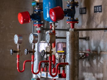 Mehrere mechanische Anzeigen im Heizkraftwerk Plauen Das Bild zeigt einen Ausschnitt eines Heizkraftwerkes. Sichtbar sind mehrere Druckanzeigen mit Zei-gern, Ventile und rote Rohrleitungen, die mit metallischen Zylindern und weiteren Komponenten ver-bunden sind.