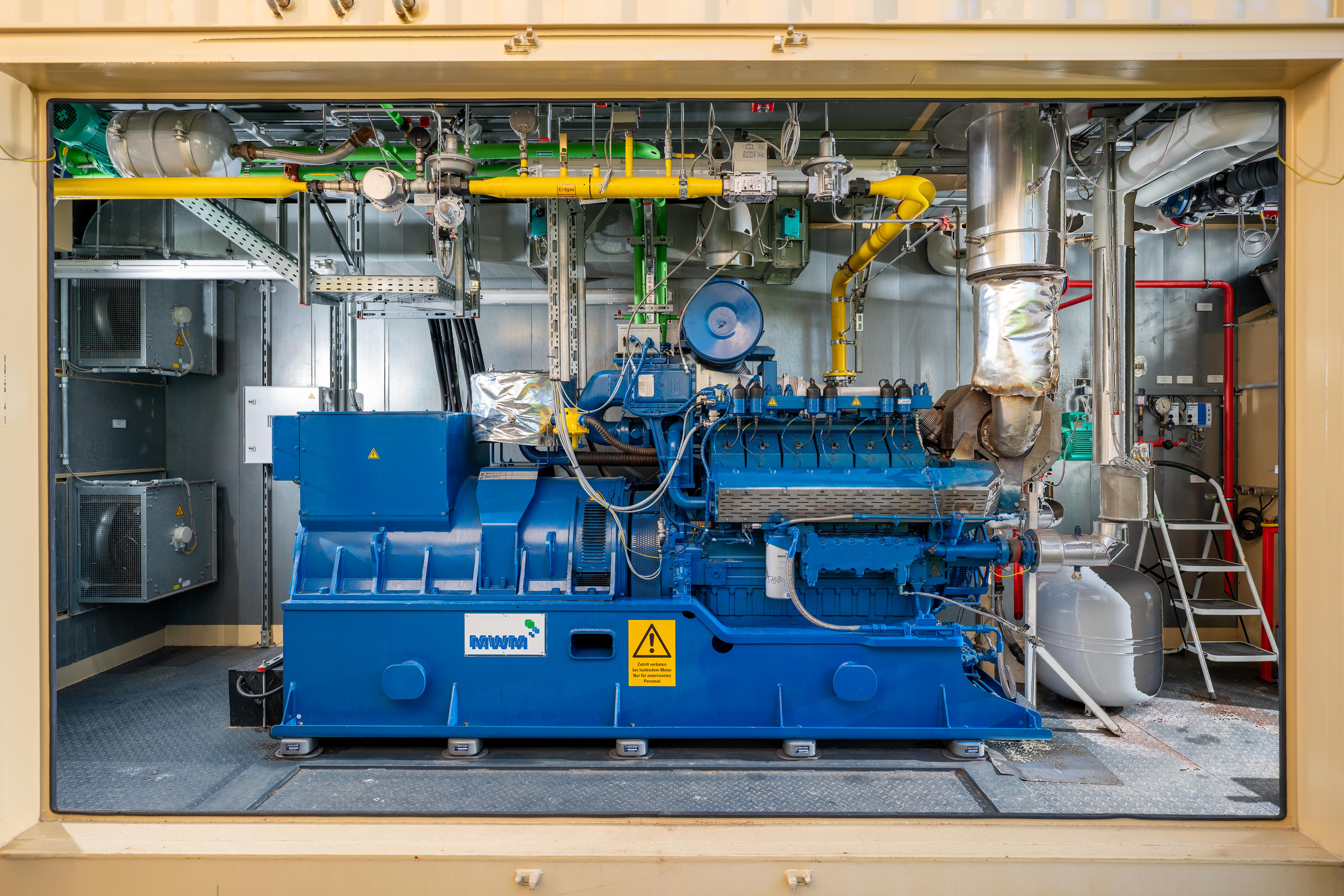 Das Bild zeigt eine blaue Industriemaschine in einer Anlage. An der Maschine sind verschiedene Rohre, Ventile und elektrische Komponenten angeschlossen. Im Hintergrund befinden sich metallene Wände und weitere Ausrüstungsgegenstände.