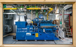 BHKW im Container Das Bild zeigt eine blaue Industriemaschine in einer Anlage. An der Maschine sind verschiedene Rohre, Ventile und elektrische Komponenten angeschlossen. Im Hintergrund befinden sich metallene Wände und weitere Ausrüstungsgegenstände.