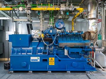 BHKW im Container Das Bild zeigt eine blaue Industriemaschine in einer Anlage. An der Maschine sind verschiedene Rohre, Ventile und elektrische Komponenten angeschlossen. Im Hintergrund befinden sich metallene Wände und weitere Ausrüstungsgegenstände.