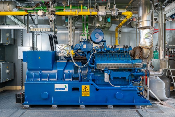 BHKW im Container Das Bild zeigt eine blaue Industriemaschine in einer Anlage. An der Maschine sind verschiedene Rohre, Ventile und elektrische Komponenten angeschlossen. Im Hintergrund befinden sich metallene Wände und weitere Ausrüstungsgegenstände.