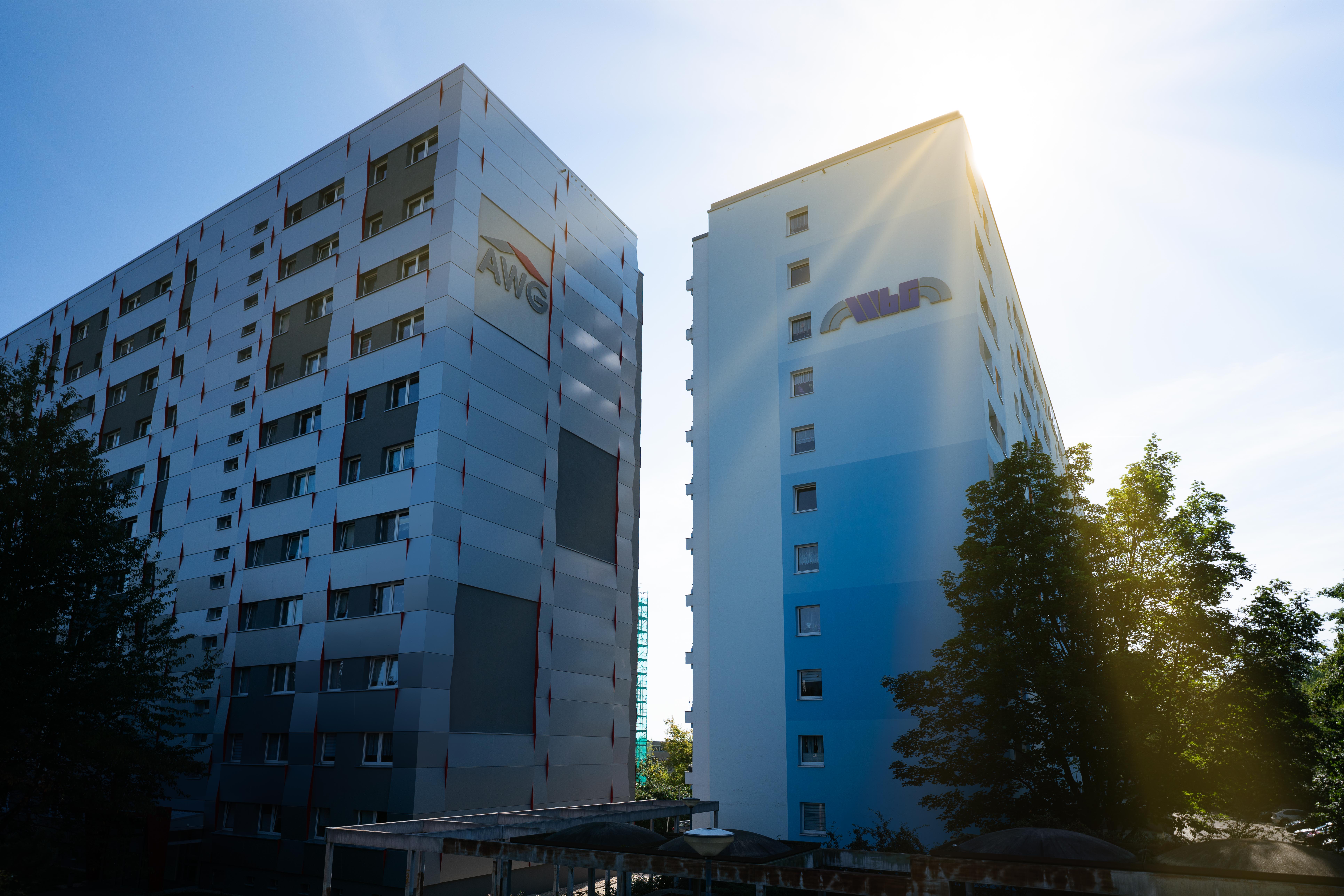 Das Bild zeigt zwei hohe Wohngebäude mit vielen Fenstern, die nebeneinander stehen. Auf beiden Ge-bäuden ist ein Logo zu erkennen. Vor den Gebäuden stehen Bäume, und der Himmel ist klar und wolkenlos.