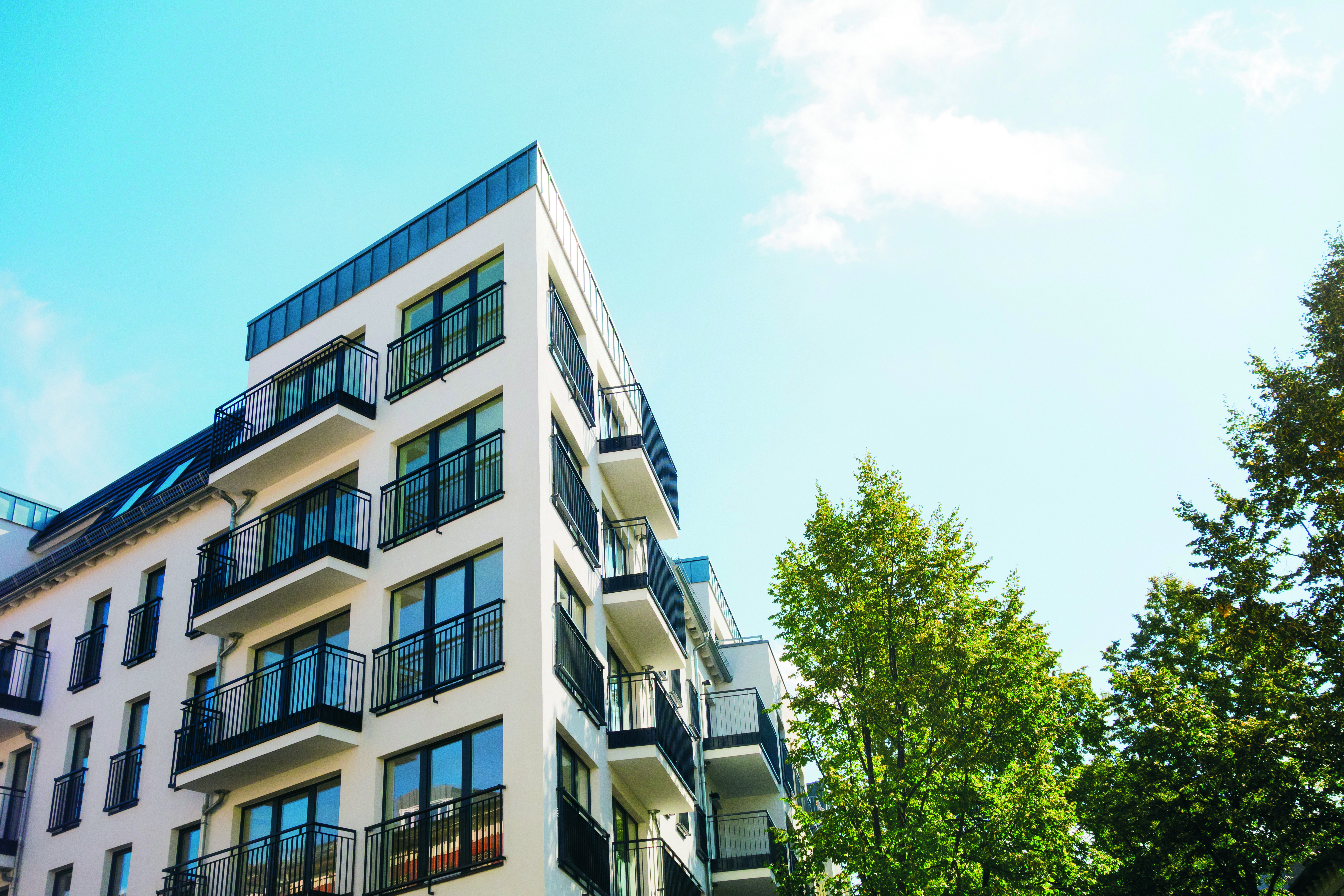 Ein modernes, mehrstöckiges Wohngebäude mit hellen Fassaden und vielen Balkonen steht vor blauem Himmel. Rechts im Bild ragt ein grüner Baum ins Bild.