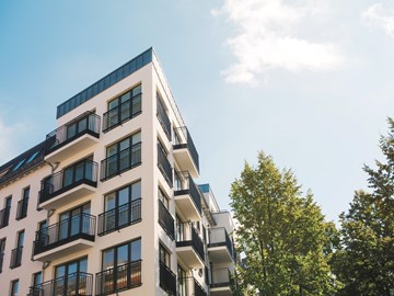 mehrstöckiges Wohngebäude Ein modernes, mehrstöckiges Wohngebäude mit hellen Fassaden und vielen Balkonen steht vor blauem Himmel. Rechts im Bild ragt ein grüner Baum ins Bild.