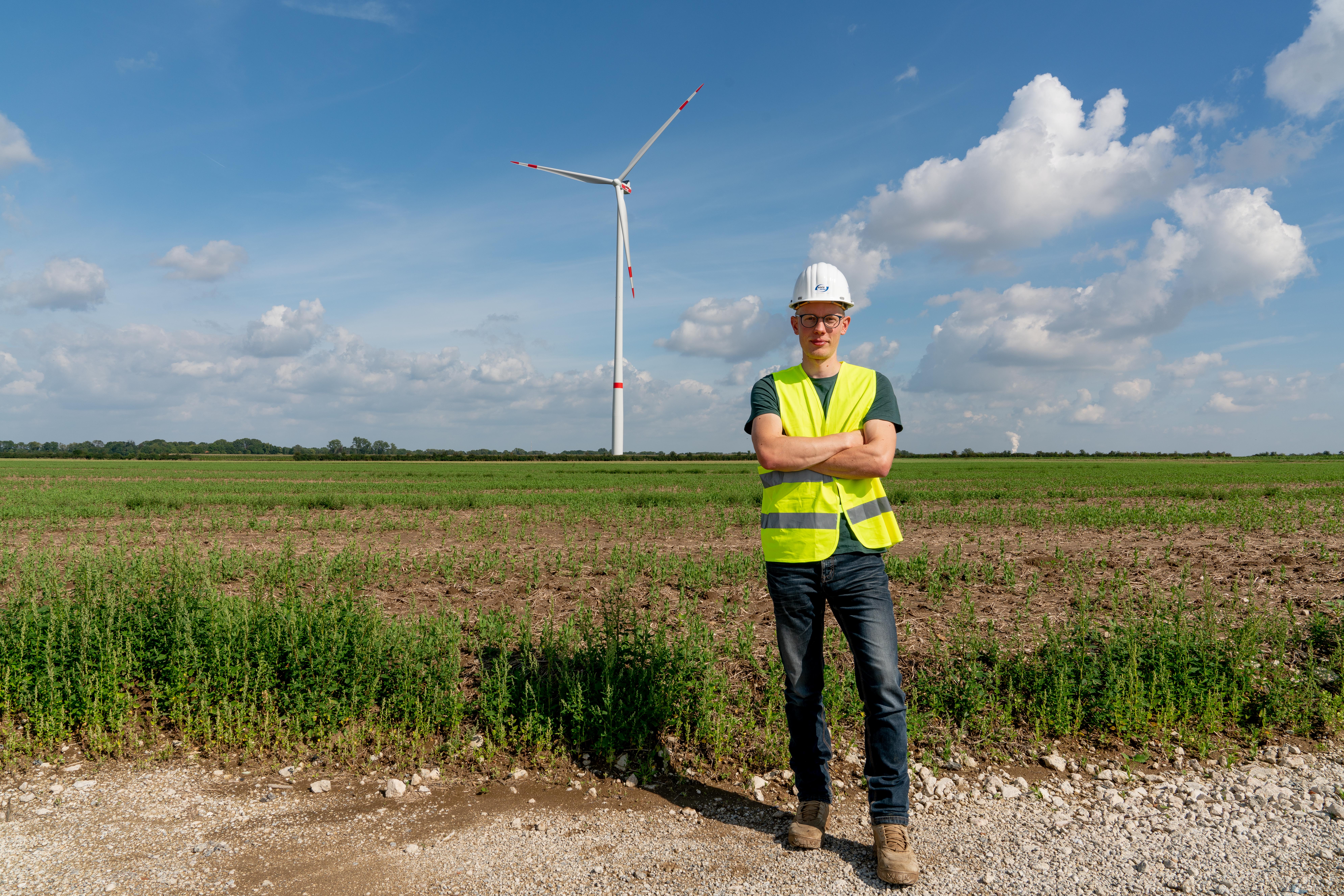 Eine Person mit Schutzhelm und gelber Warnweste steht mit verschränkten Armen auf einem Feldweg. Hinter ihr erstreckt sich ein großes grünes Feld mit einem einzelnen Windrad unter blauem Himmel mit Wolken.