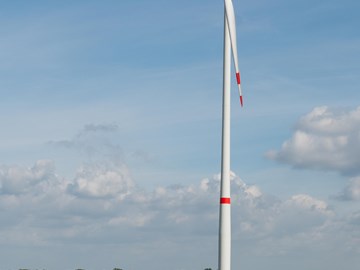 Mitarbeiter vor einer Windkraftanlage Eine Person mit Schutzhelm und gelber Warnweste steht mit verschränkten Armen auf einem Feldweg. Hinter ihr erstreckt sich ein großes grünes Feld mit einem einzelnen Windrad unter blauem Himmel mit Wolken.