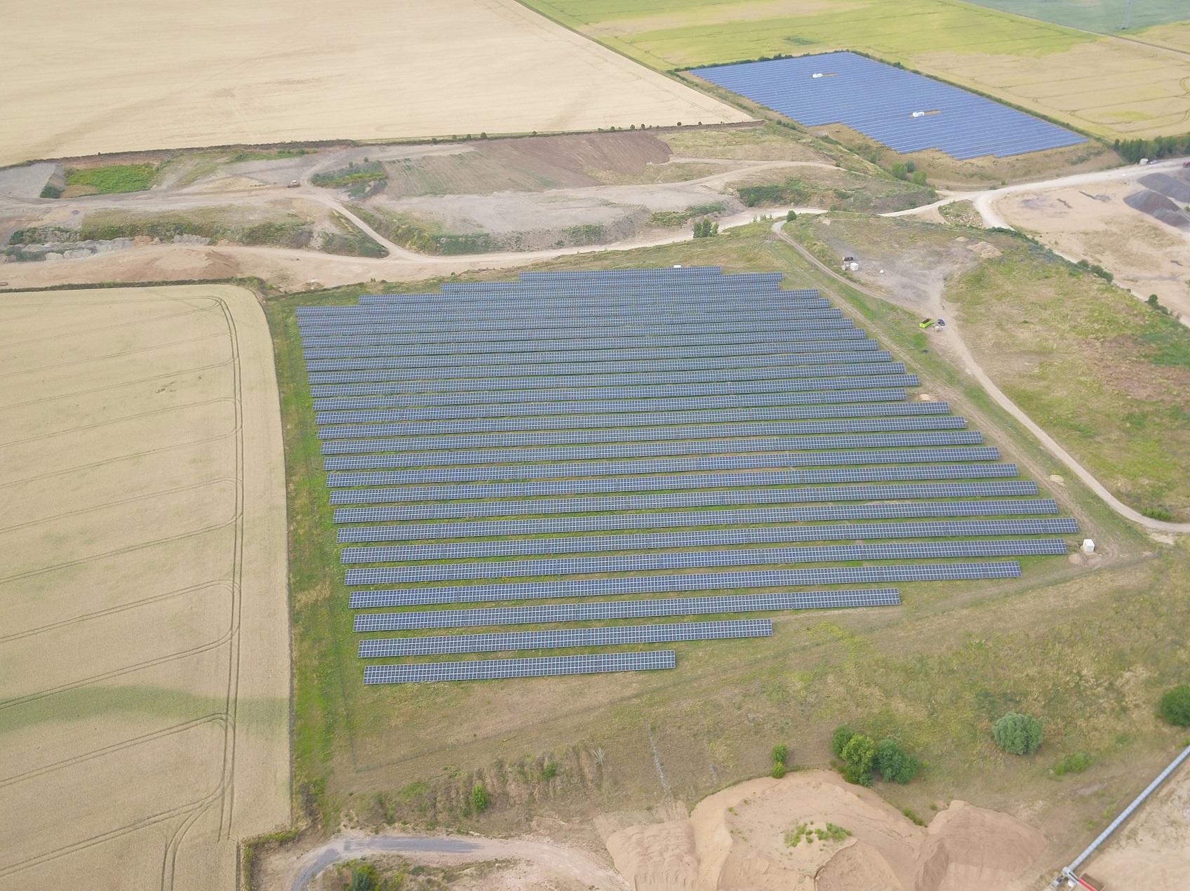 Das Bild zeigt eine Luftaufnahme eines Solarparks. Mehrere Reihen von Solarpaneelen sind auf einem weitläufigen Feld installiert. Umgeben ist die Anlage von weiteren Feldern, teils landwirtschaftlich genutzt.