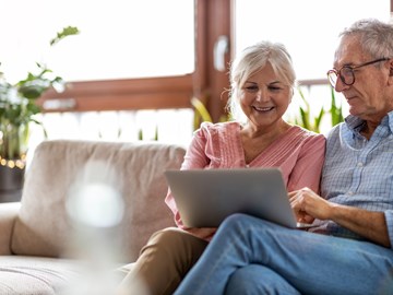 Frau und Mann schauen auf dem Laptop in das Kundenportal Frau und Mann sitzen auf dem Sofa vor dem Fenster und schauen auf dem Laptop in das Kundenportal