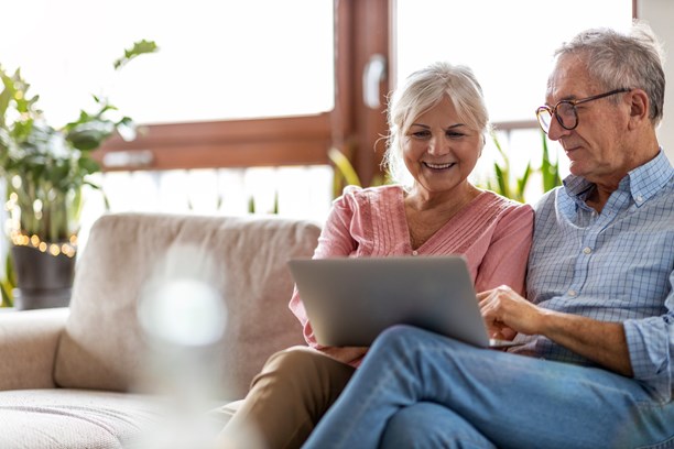 Frau und Mann schauen auf dem Laptop in das Kundenportal Frau und Mann sitzen auf dem Sofa vor dem Fenster und schauen auf dem Laptop in das Kundenportal