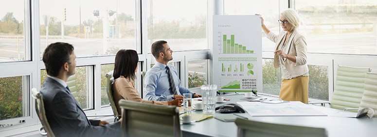 Frau erläutert einer Frau und einem Mann Diagramme auf Flip-Chart bei Konferenz Frau erläutert einer Frau und einem Mann Diagramme auf Flip-Chart bei Konferenz