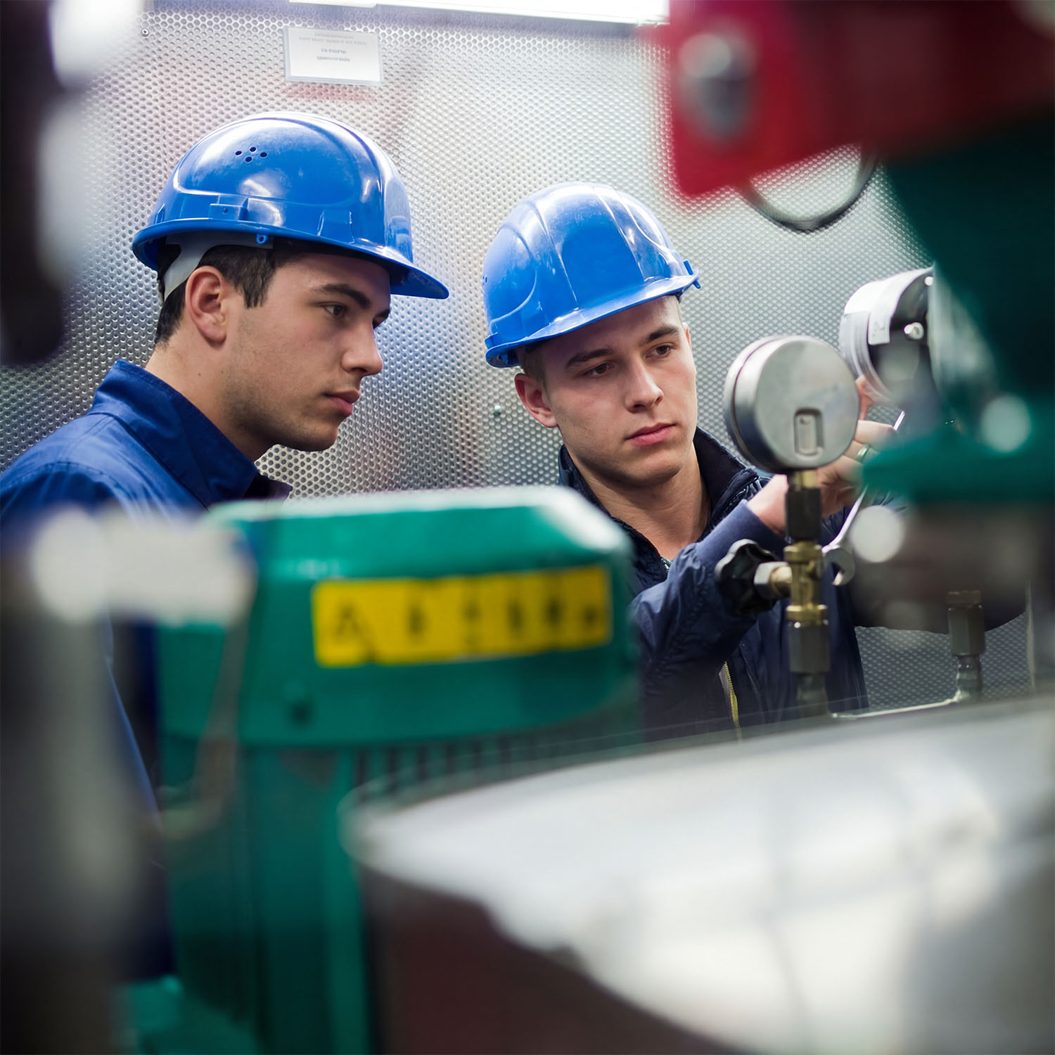 Zwei Personen mit blauen Schutzhelmen und dunkelblauer Arbeitskleidung arbeiten in einem technischen Raum. Sie bedienen Messgeräte und Ventile an einer Maschine. Im Vordergrund sind verschiedene Rohre und Geräte zu sehen.
