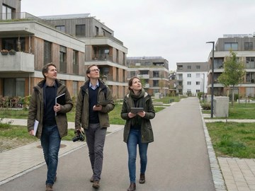 Bild KI generiert: Drei Personen in einem Wohngebiet Drei Personen gehen eine breite Fußgängerallee in einem modernen Wohngebiet entlang. Sie tragen Jacken und halten Unterlagen oder Tablets in den Händen. Rechts und links stehen mehrstöckige Wohngebäude mit Balkonen. Der Himmel ist bedeckt.