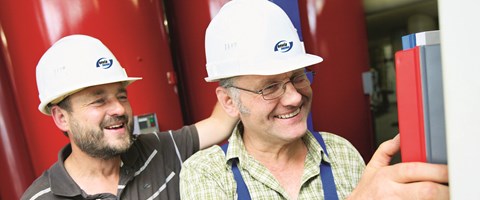 Nahaufnahme, zwei Männer mit envia THERM-Schutzhelmen stehen lachend vor roten Anlagenteilen Nahaufnahme, zwei Männer mit envia THERM-Schutzhelmen stehen lachend vor roten Anlagenteilen