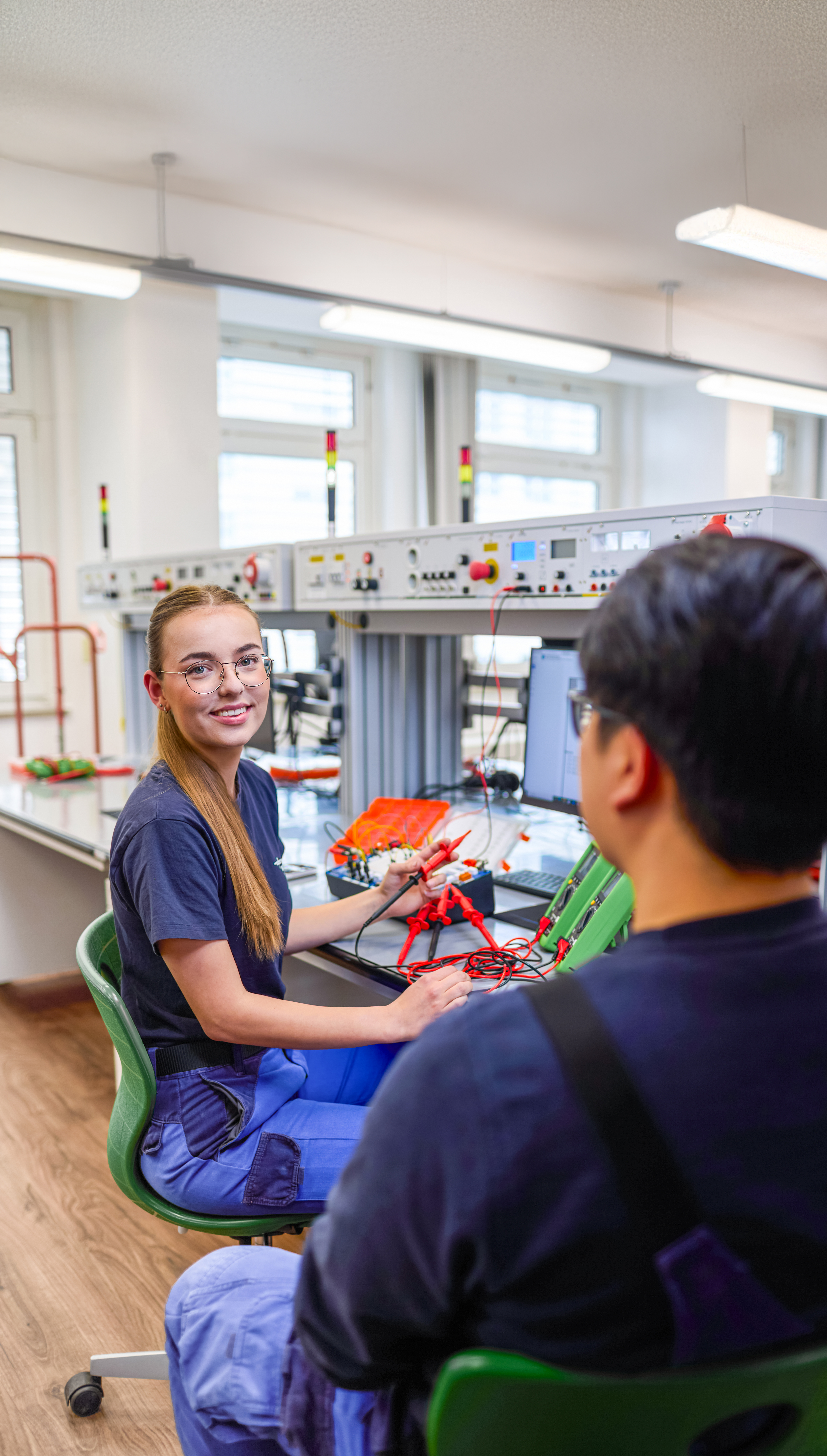 Zwei Personen sitzen in einem Werkraum an einem Arbeitstisch und arbeiten mit elektronischen Bauteilen und Messgeräten. Auf dem Tisch liegen Kabel, Geräte und Werkzeuge. Im Hintergrund sind weitere Arbeitsplätze mit technischen Anlagen zu sehen.