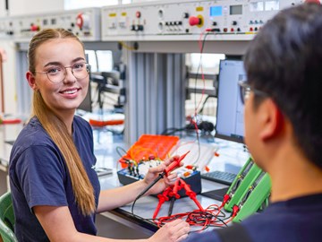 Ausbildung zur Elektronikerin für Betriebstechnik Zwei Personen sitzen in einem Werkraum an einem Arbeitstisch und arbeiten mit elektronischen Bauteilen und Messgeräten. Auf dem Tisch liegen Kabel, Geräte und Werkzeuge. Im Hintergrund sind weitere Arbeitsplätze mit technischen Anlagen zu sehen.