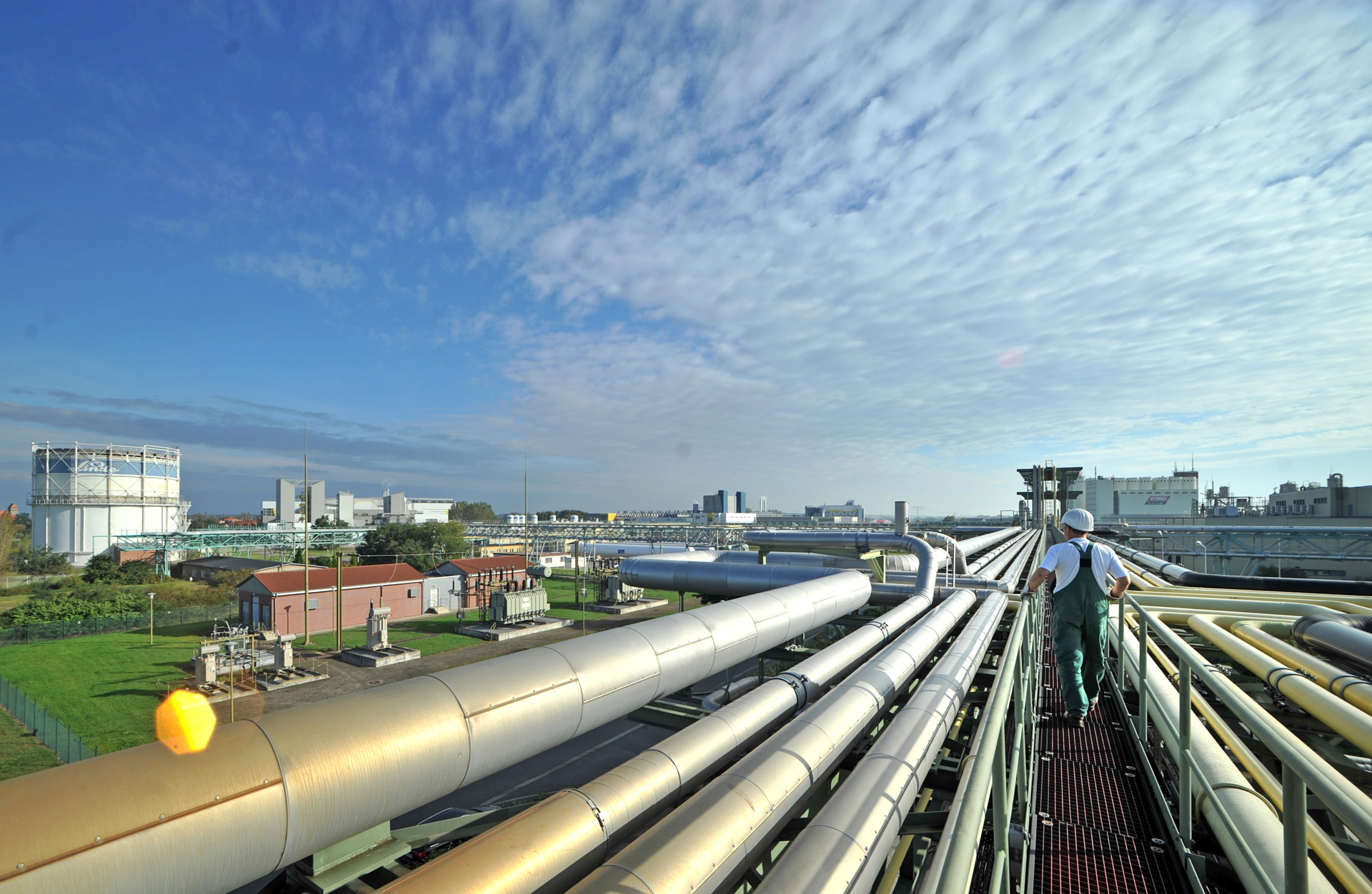 Das Foto zeigt eine industrielle Anlage mit zahlreichen großen Rohrleitungen, die parallel zueinander verlaufen. Zwischen den Rohren befindet sich ein schmaler Laufsteg, auf dem eine Person in Schutzkleidung und mit Helm entlanggeht.