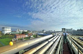 Rohrbrücken im Chemiepark Bitterfeld © Chemiepark Bitterfeld-Wolfen GmbH, Fotograf: André Kehrer Das Foto zeigt eine industrielle Anlage mit zahlreichen großen Rohrleitungen, die parallel zueinander verlaufen. Zwischen den Rohren befindet sich ein schmaler Laufsteg, auf dem eine Person in Schutzkleidung und mit Helm entlanggeht.