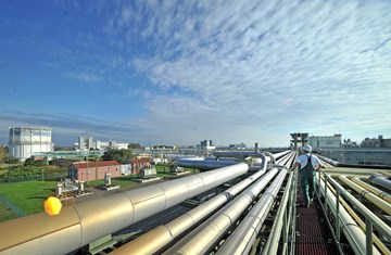 Rohrbrücken im Chemiepark Bitterfeld © Chemiepark Bitterfeld-Wolfen GmbH, Fotograf: André Kehrer Das Foto zeigt eine industrielle Anlage mit zahlreichen großen Rohrleitungen, die parallel zueinander verlaufen. Zwischen den Rohren befindet sich ein schmaler Laufsteg, auf dem eine Person in Schutzkleidung und mit Helm entlanggeht.