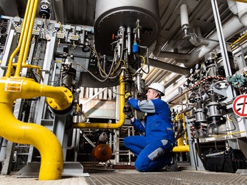 Mechatroniker bei envia THERM Das Foto zeigt eine Person in blauer Arbeitskleidung und mit weißem Schutzhelm, die Wartungs- oder Inspektionsarbeiten an einer industriellen Anlage durchführt. Die Umgebung besteht aus verschiedenen Rohren, Ventilen und technischen Geräten.