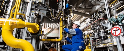 Mechatroniker bei envia THERM Das Foto zeigt eine Person in blauer Arbeitskleidung und mit weißem Schutzhelm, die Wartungs- oder Inspektionsarbeiten an einer industriellen Anlage durchführt. Die Umgebung besteht aus verschiedenen Rohren, Ventilen und technischen Geräten.