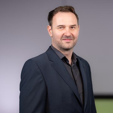 Norman Driebe Eine Person steht vor einem neutralen Hintergrund. Sie trägt einen dunkelblauen Anzug und ein dunkles Hemd.