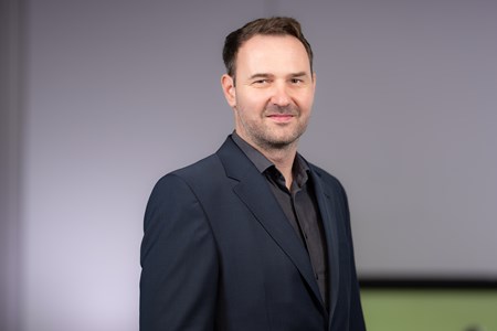 Norman Driebe Eine Person steht vor einem neutralen Hintergrund. Sie trägt einen dunkelblauen Anzug und ein dunkles Hemd.