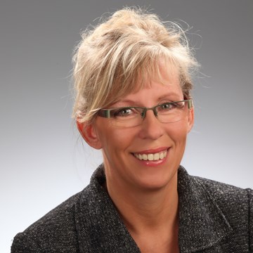 Ute Haupt Eine Person mit nach hinten gebundenen blonden Haaren ist zu sehen. Sie trägt eine Brille und ein dunkel gefärbtes Sakko.