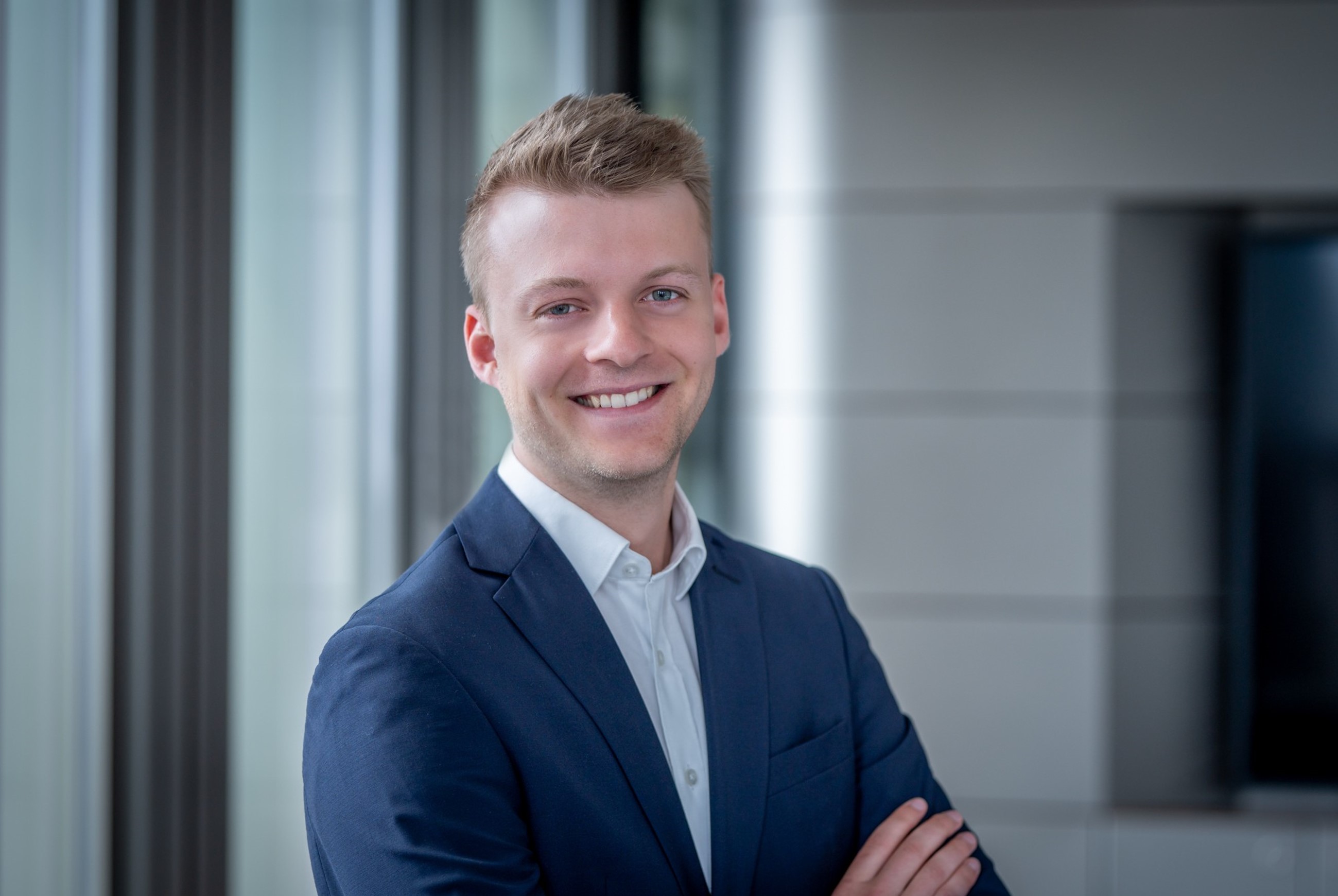 Eine Person steht mit verschränkten Armen. Sie trägt einen blauen Anzug und ein weißes Hemd. Im Hintergrund sind vertikale Fensterjalousien sowie ein Wandabschnitt mit horizontalen Linien zu sehen.