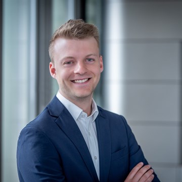 Justus Nowak Eine Person steht mit verschränkten Armen. Sie trägt einen blauen Anzug und ein weißes Hemd. Im Hintergrund sind vertikale Fensterjalousien sowie ein Wandabschnitt mit horizontalen Linien zu sehen.