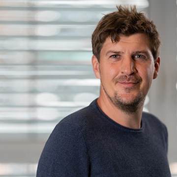 Peter Duever Eine Person steht vor einem Fenster mit horizontalen Jalousien. Die Person trägt einen dunkelblauen Pullover.