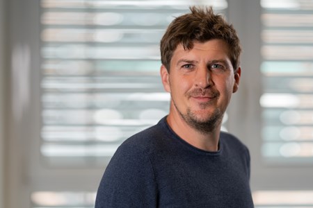 Peter Duever Eine Person steht vor einem Fenster mit horizontalen Jalousien. Die Person trägt einen dunkelblauen Pullover.