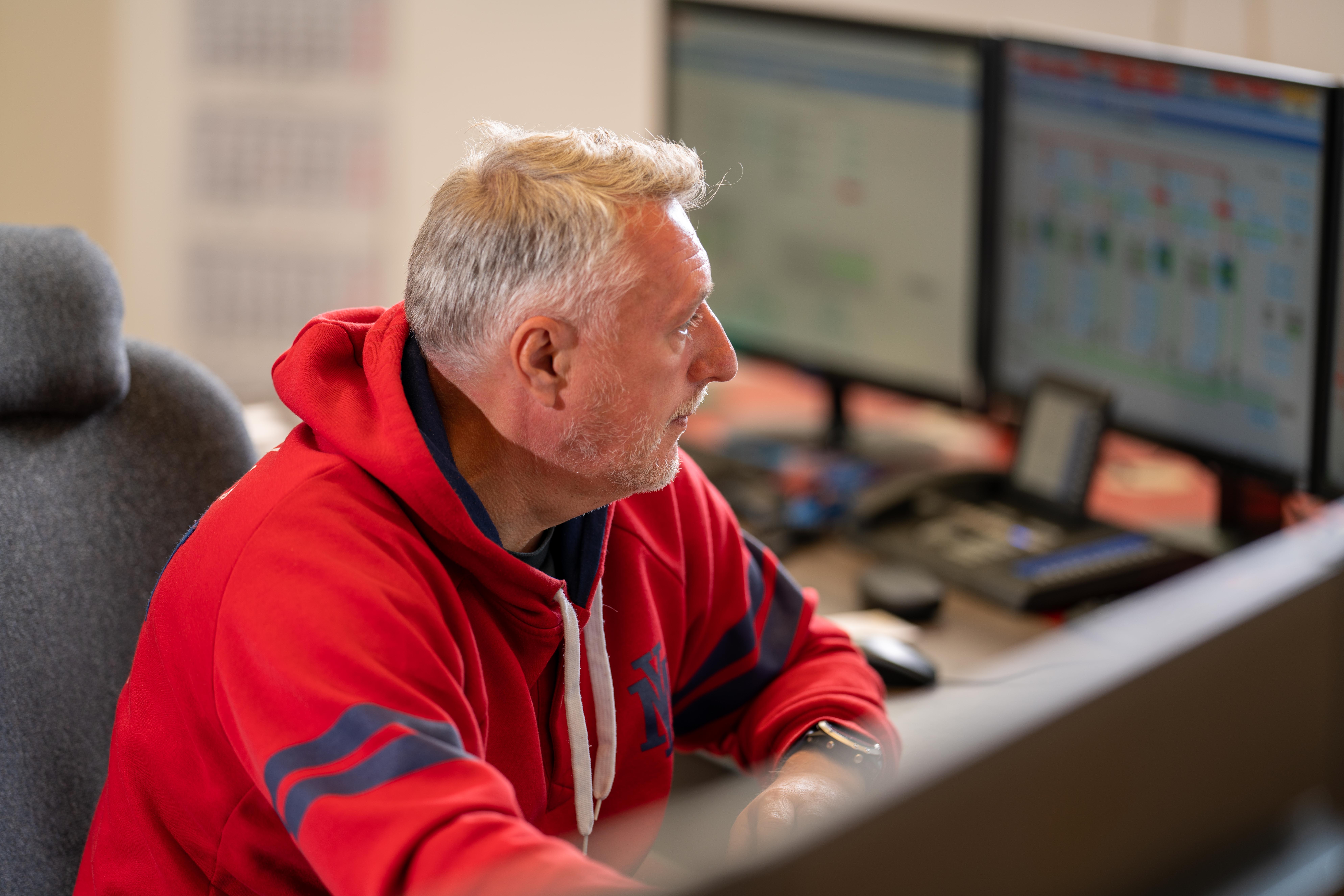 Eine Person sitzt vor mehreren Computerbildschirmen. Sie trägt einen roten Hoodie mit blauen Akzenten. Der Hintergrund ist leicht unscharf, wodurch der Fokus auf der Person und der Computeranordnung liegt.