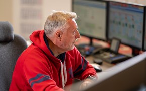 Stoerungsmeldung Eine Person sitzt vor mehreren Computerbildschirmen. Sie trägt einen roten Hoodie mit blauen Akzenten. Der Hintergrund ist leicht unscharf, wodurch der Fokus auf der Person und der Computeranordnung liegt.