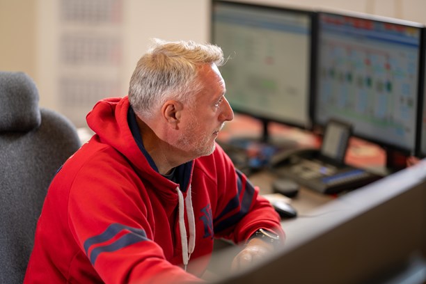 Stoerungsmeldung Eine Person sitzt vor mehreren Computerbildschirmen. Sie trägt einen roten Hoodie mit blauen Akzenten. Der Hintergrund ist leicht unscharf, wodurch der Fokus auf der Person und der Computeranordnung liegt.