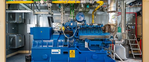 BHKW in Container Das Bild zeigt eine blaue Industriemaschine in einer Anlage. An der Maschine sind verschiedene Rohre, Ventile und elektrische Komponenten angeschlossen. Im Hintergrund befinden sich metallene Wände und weitere Ausrüstungsgegenstände.