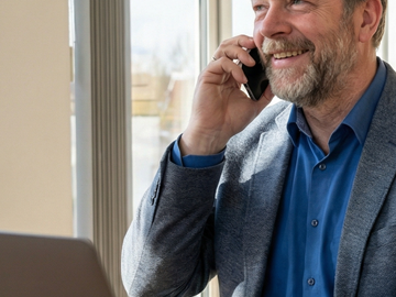 Mann sitzt telefonierend an einem Tisch vor einem Laptop Eine Person sitzt an einem Holztisch in einem hellen Büro und telefoniert, während sie an einem Laptop arbeitet. Große Fenster lassen viel Tageslicht herein, im Hintergrund sind moderne Wandobjekte und eine Zimmerpflanze zu sehen.