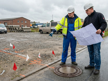Zwei Personen mit Schutzhelmen stehen auf einer Baustelle. Zwei Personen mit Schutzhelmen stehen auf einer Baustelle. Eine Person zeigt auf den Boden, die andere hält einen Bauplan in den Händen. Auf dem Gelände sind Markierungen, Absperrfähnchen, Fahrzeuge und Baumaterialien zu sehen.